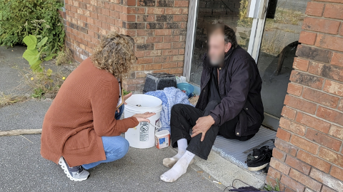 Kneeling SW Man Socks A Community Care social worker speaking with a man sitting outside a building.